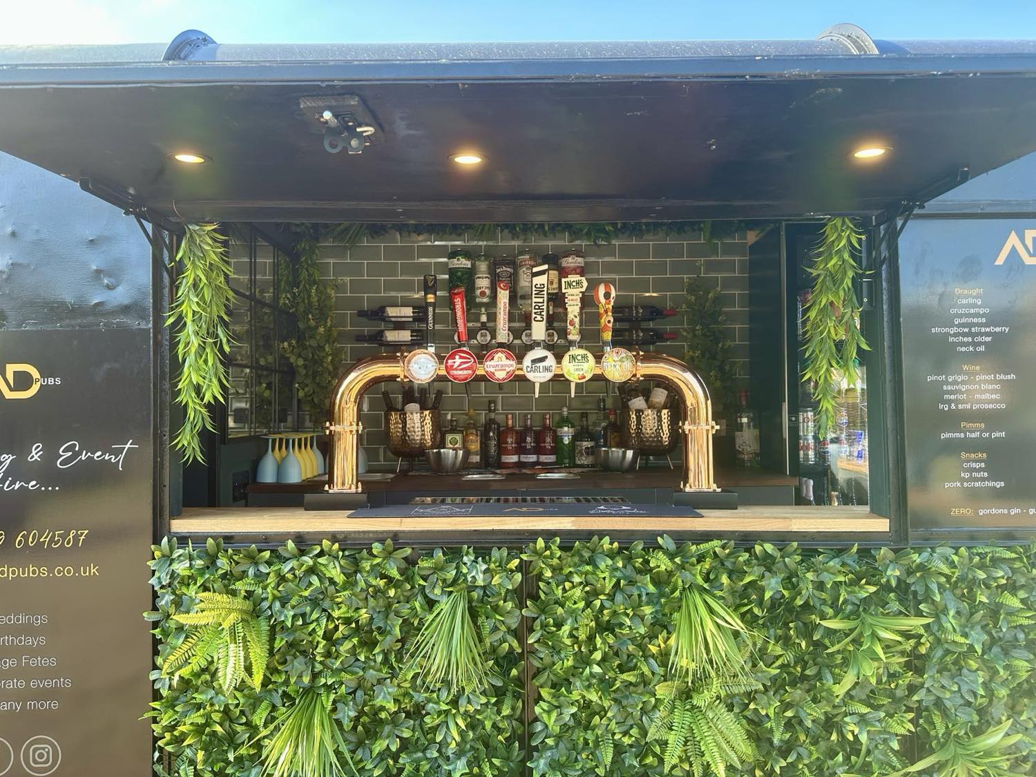 The ADevents horsebox bar front-on - copper taps, foliage and the drinks menu.
