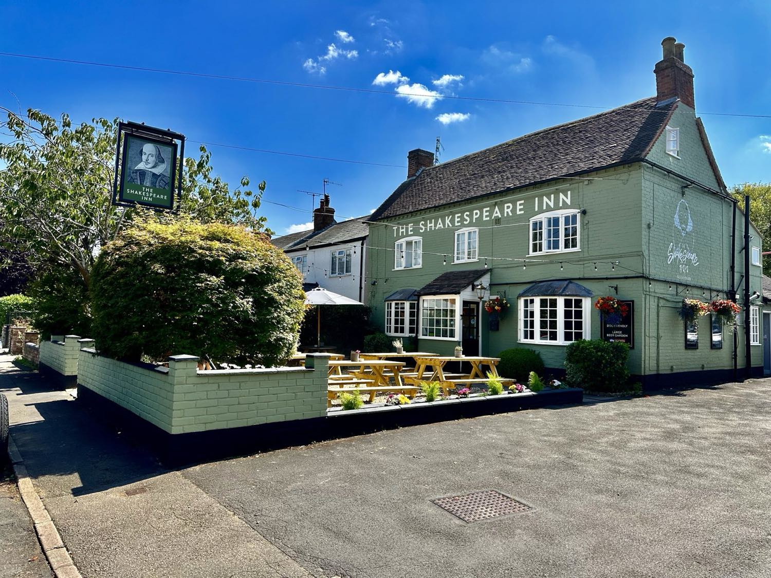 The Shakespeare Inn - a sage-green painted village pub under a blue sky, with its hanging pub sign and picnic benches out front.